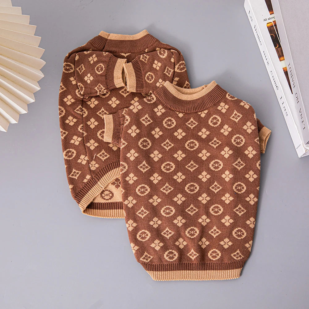 Two brown patterned dog sweaters on a gray surface with books and a fan in the background.